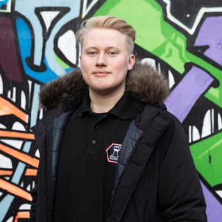 A University of Bradford student wearing a black coat stood in front of a colourful graffiti wall.