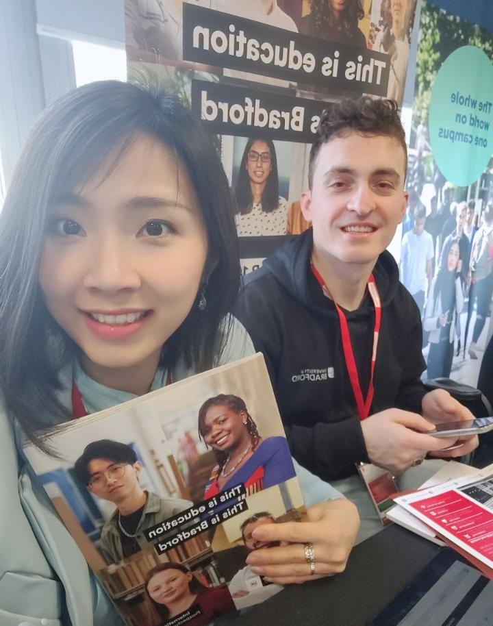 Annie Pham at a recruitment fair for international students holding a copy of the international prospectus. Next to her is an international student ambassador in front of a display board advertising the University of Bradford
