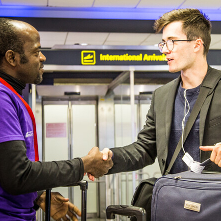 Student holding luggage and shaking the hand of a member of our International Support team in an Airport. The background shows a sign that says International Arrivals.