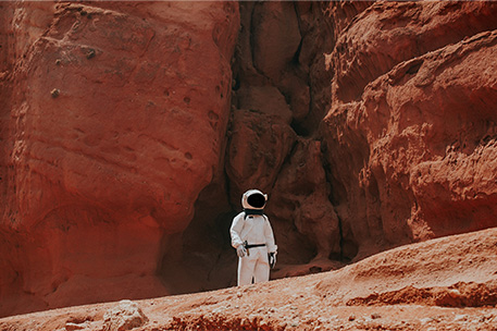 A person wearing a space suit on a dessert landscape with red cliffes in the background. The landscape represents Mars.