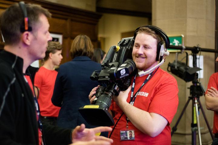 A smiling male student operating a television camera and wearing a BBC t-shirt 