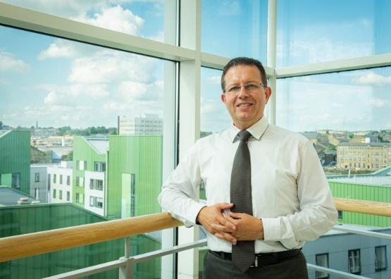 A university staff member wearing a shirt and tie is stood up in front of a glass window in a building A university staff member wearing a shirt and tie is stood up in front of a glass window in a building