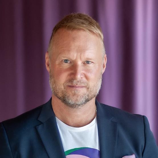 A head and shoulders photograph of a person wearing a T-shirt and jacket poses in front of a purple coloured curtain A head and shoulders photograph of a person wearing a T-shirt and jacket poses in front of a purple coloured curtain