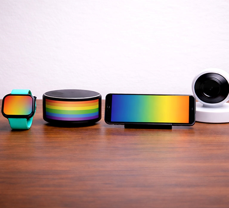 A smartwatch, smart speaker, smartphone and security camera arranged on a wooden table, each shown with rainbow-coloured display elements against a plain white background.