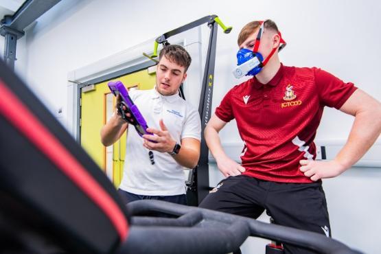 A football player wearing a tracksuit on an exercise bike while wearing a face mask gets a demonstration from a student holding a tablet computer A football player wearing a tracksuit on an exercise bike while wearing a face mask gets a demonstration from a student holding a tablet computer