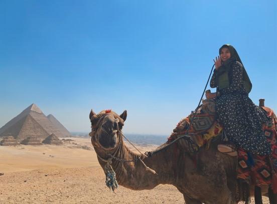 Jubeda Farzi riding a camel with pyramids in the background Jubeda Farzi riding a camel with pyramids in the background