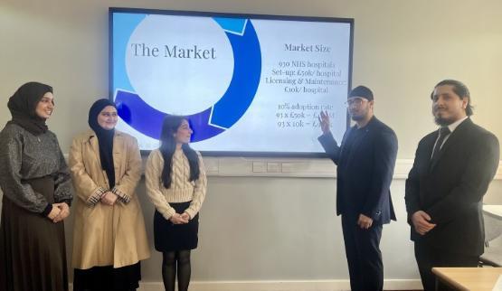 THree students stood on the left and another two students stood on the right present a pitch using a projector screen on a wall behind them THree students stood on the left and another two students stood on the right present a pitch using a projector screen on a wall behind them