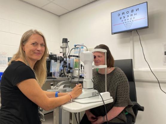 A person on the left in charge of a mock eye test as another person leans forward on testing equipment A person on the left in charge of a mock eye test as another person leans forward on testing equipment