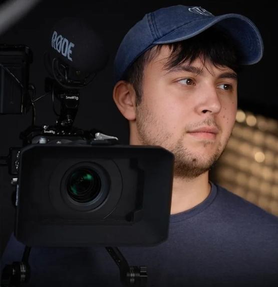 former student wearing baseball cap holding a TV camera over their right shoulder former student wearing baseball cap holding a TV camera over their right shoulder
