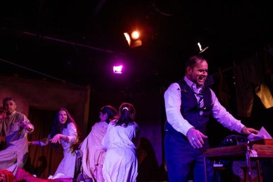 A person stood up dressed in a waistcoat and tie cries out while a group of four people do a dance and snarl towards the person A person stood up dressed in a waistcoat and tie cries out while a group of four people do a dance and snarl towards the person