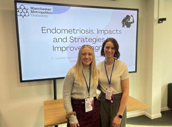 Two people wearing lanyards stand next to each other and smile in front of a screen after giving a talk Two people wearing lanyards stand next to each other and smile in front of a screen after giving a talk