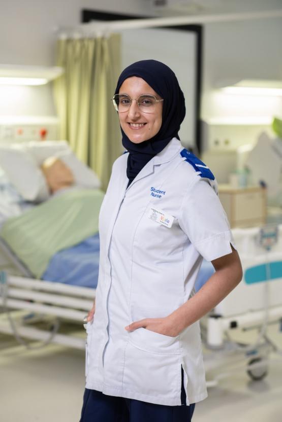 A university student stands up in front of a bed in a mock hospital ward wearing a uniform with student nurse written on it A university student stands up in front of a bed in a mock hospital ward wearing a uniform with student nurse written on it