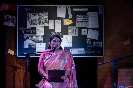 A person sat down in a sequin covered dress speaking behind a microphone in front of a board covered in newspaper cutouts A person sat down in a sequin covered dress speaking behind a microphone in front of a board covered in newspaper cutouts