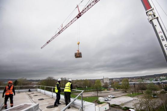 Two people on a roof watch as a crane above them delivers a large wooden box onto the roof Two people on a roof watch as a crane above them delivers a large wooden box onto the roof