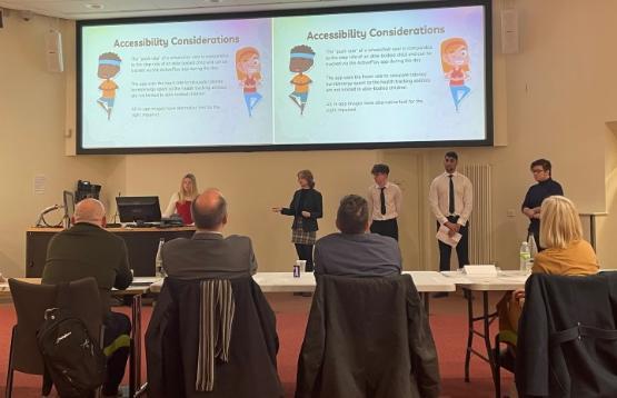 A group of students in the background stood up delivering a pitch to people sat at a table in the picture's foreground A group of students in the background stood up delivering a pitch to people sat at a table in the picture's foreground