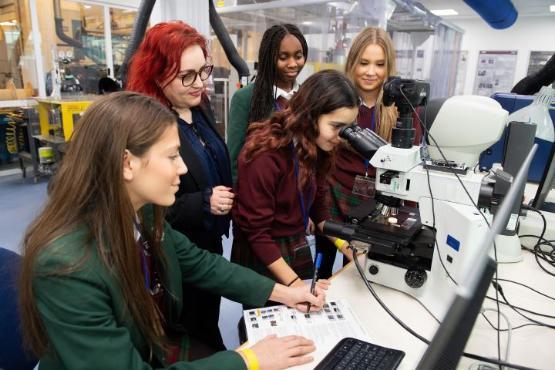 Four students and a staff member stood around a microscope in an engineering room Four students and a staff member stood around a microscope in an engineering room