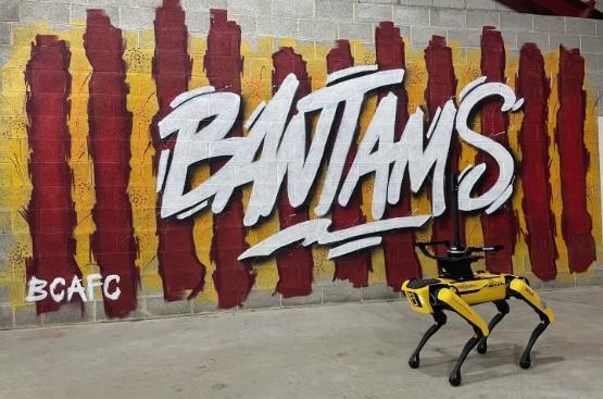 A robot dog to the right of a photo in front of a wall with claret and amber coloured street art with Bantams written on it A robot dog to the right of a photo in front of a wall with claret and amber coloured street art with Bantams written on it
