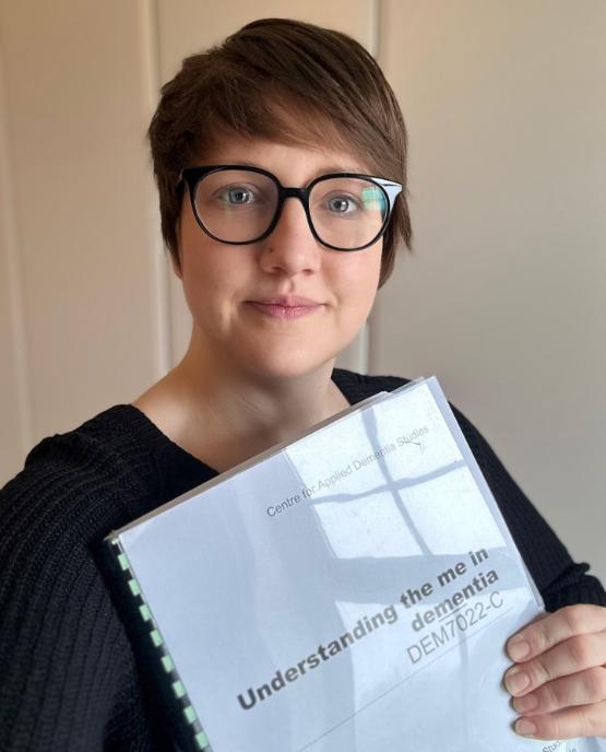 A person wearing glasses stood up holding an A4-sized paper document close to them A person wearing glasses stood up holding an A4-sized paper document close to them