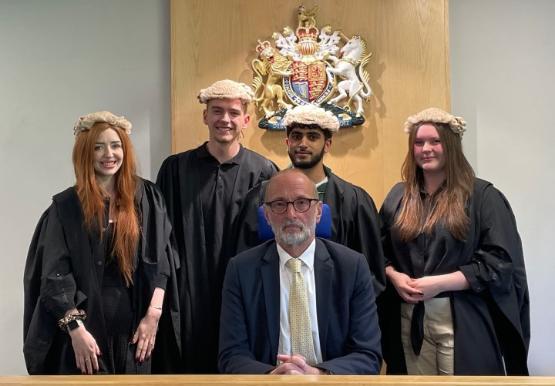 A teacher and school students in legal ropes and wigs stood up behind a seated judge A teacher and school students in legal ropes and wigs stood up behind a seated judge