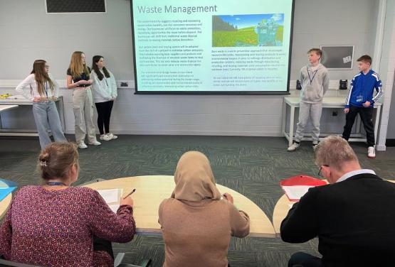 Five students stood either side of a projectrion screen give a pitch to a group of judges sat at a table Five students stood either side of a projectrion screen give a pitch to a group of judges sat at a table