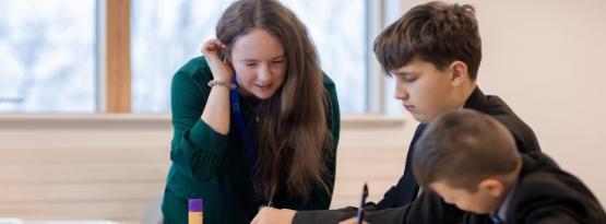 A teacher stands over between two school pupils who are sat at a wooden desk A teacher stands over between two school pupils who are sat at a wooden desk