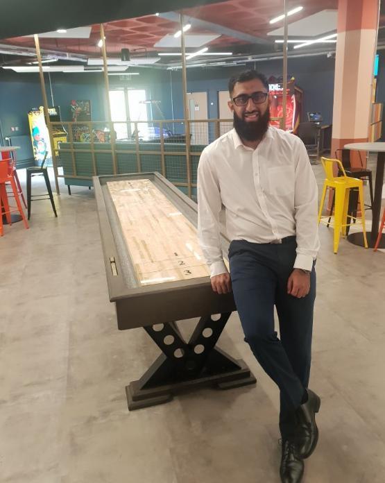 A university staff member stands to the side of a wooden shuffleboard table inside a students' union room A university staff member stands to the side of a wooden shuffleboard table inside a students' union room