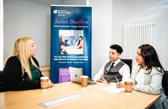 Three people sat at wooden table talking with Justice Bradford banner in background Three people sat at wooden table talking with Justice Bradford banner in background