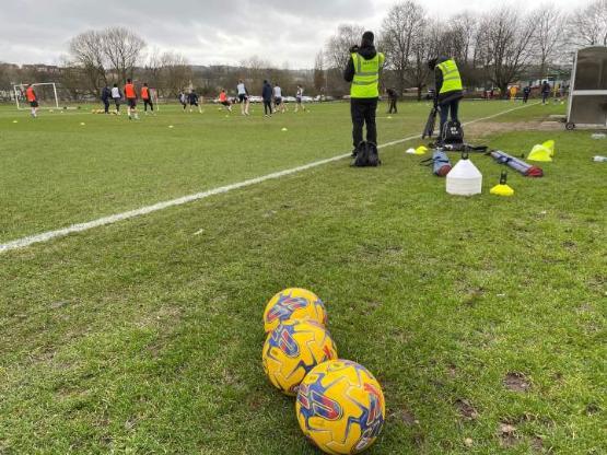 two students in hi-vis jackets filming football training session two students in hi-vis jackets filming football training session
