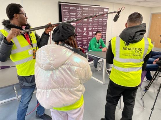 A group of students film a press conference with a football player sat down at a desk A group of students film a press conference with a football player sat down at a desk