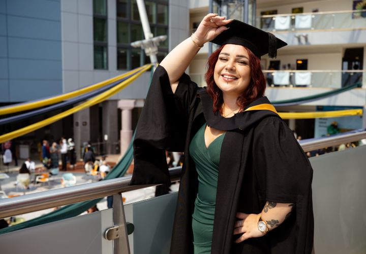 A student dressed in graduation gown holds their cap by one hand A student dressed in graduation gown holds their cap by one hand