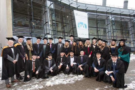 A large group of students dressed in graduation gowns pose for photo outside university building A large group of students dressed in graduation gowns pose for photo outside university building