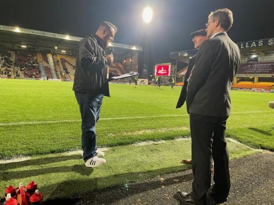 A person holding a microphone stands to the side of two university staff members who are being interviewed on the touchline at a football stadium A person holding a microphone stands to the side of two university staff members who are being interviewed on the touchline at a football stadium