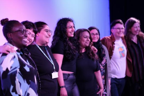 A group of six people stand up next to each other on a stage at a show and all smile A group of six people stand up next to each other on a stage at a show and all smile