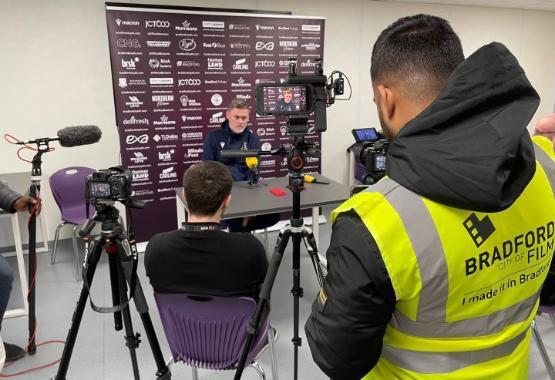 a student stands behind video camera on a tripod filming a press conference with a manager sitting at a desk a student stands behind video camera on a tripod filming a press conference with a manager sitting at a desk