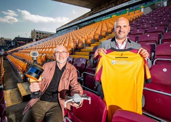Two people sit next to each other in an empty football stadium's stand, one of them holds a drone while the other person holds a football shirt in front of them Two people sit next to each other in an empty football stadium's stand, one of them holds a drone while the other person holds a football shirt in front of them