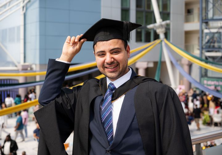 A student dressed in a suit and graduation gown and cap poses for a photo