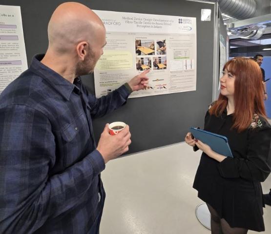 A person holding a coffee points at a poster while a person stood opposite them looks on A person holding a coffee points at a poster while a person stood opposite them looks on