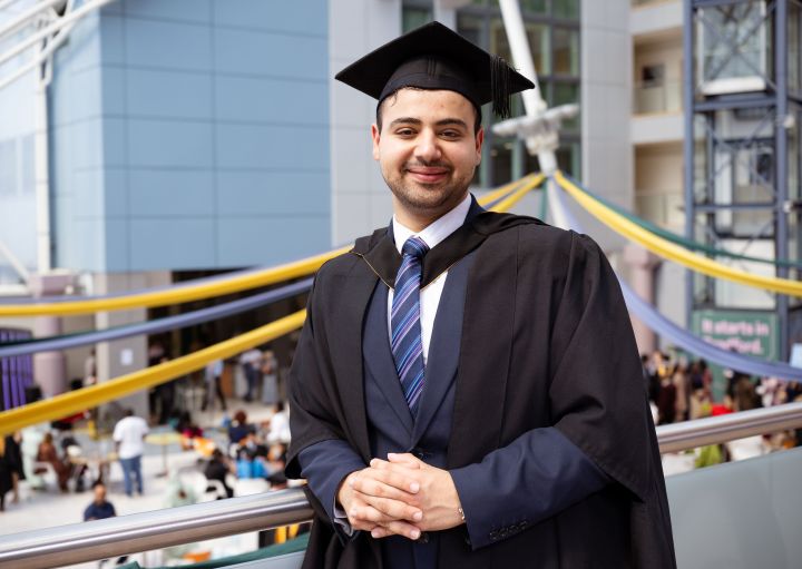 A student dressed in a graduation cap and gown leans against a balcony rail A student dressed in a graduation cap and gown leans against a balcony rail