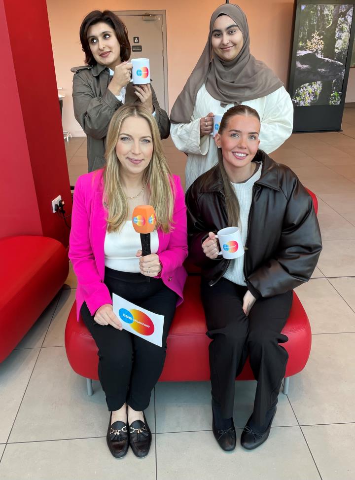 Two people holding mugs stand up behind two other people sat on a sofa in front of them Two people holding mugs stand up behind two other people sat on a sofa in front of them