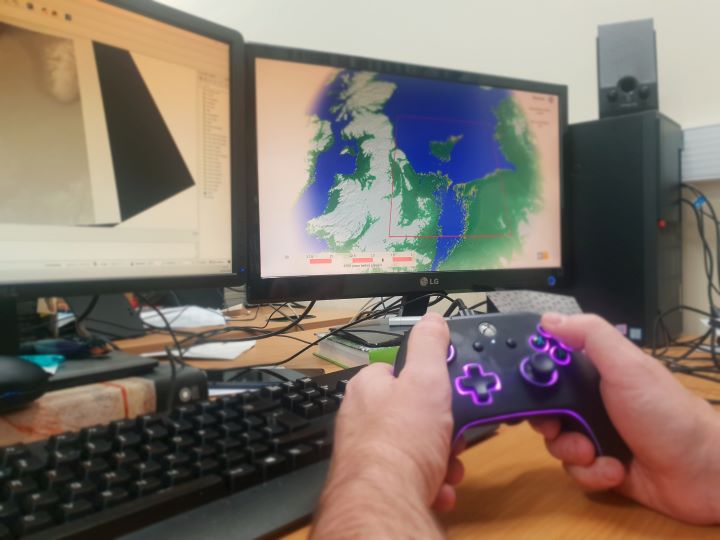 A person using a video games console controller wit two computer screens on a table in front of them A person using a video games console controller wit two computer screens on a table in front of them