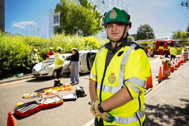 A close-up picture of a paramedic student in uniform with a crash scene behind them A close-up picture of a paramedic student in uniform with a crash scene behind them