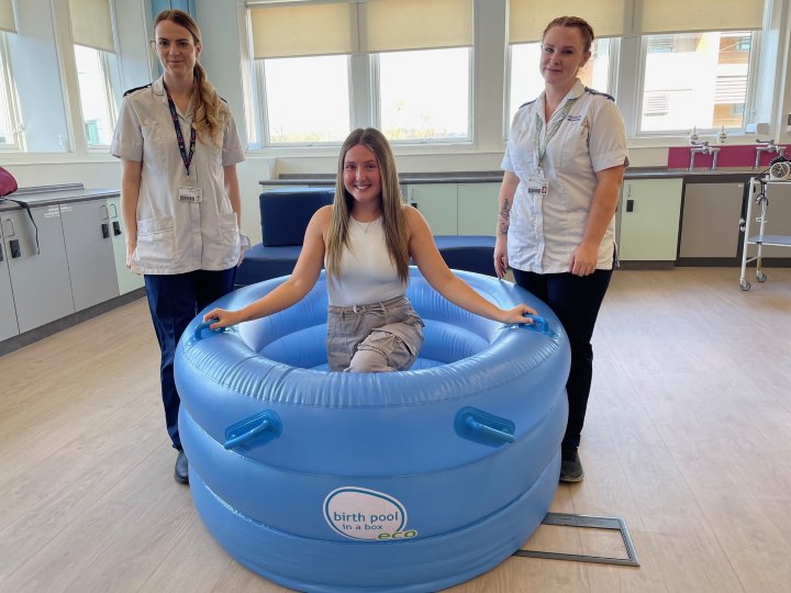Two students dressed in white midwife uniforms stand outside an inflatable birthing pool between them that is empty and has a third person in it Two students dressed in white midwife uniforms stand outside an inflatable birthing pool between them that is empty and has a third person in it