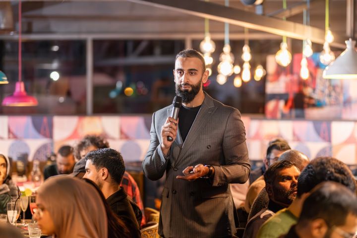 A person dressed in a business suit holds a microphone and talks in between tables of diners in a restaurant A person dressed in a business suit holds a microphone and talks in between tables of diners in a restaurant