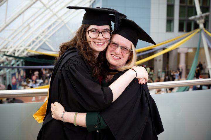 A mother and daughter hug dressed in graduation gowns