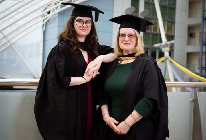 A mother and daughter stand next to each other in graduation gowns A mother and daughter stand next to each other in graduation gowns
