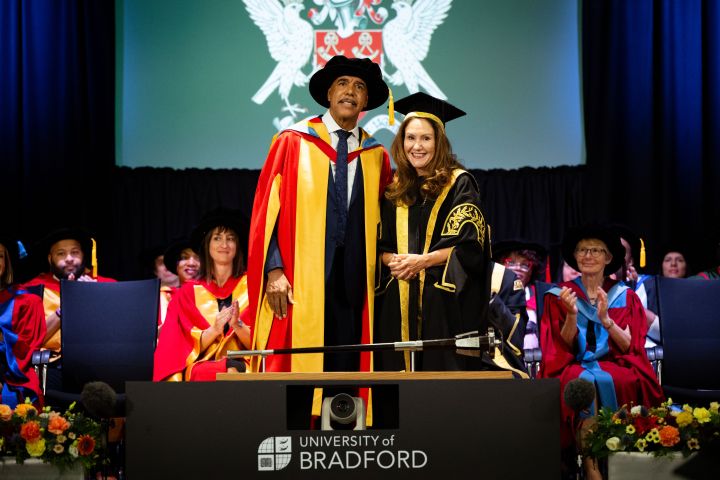 Two people dressed in graduation gowns stand next to each other on a ceremony stage Two people dressed in graduation gowns stand next to each other on a ceremony stage