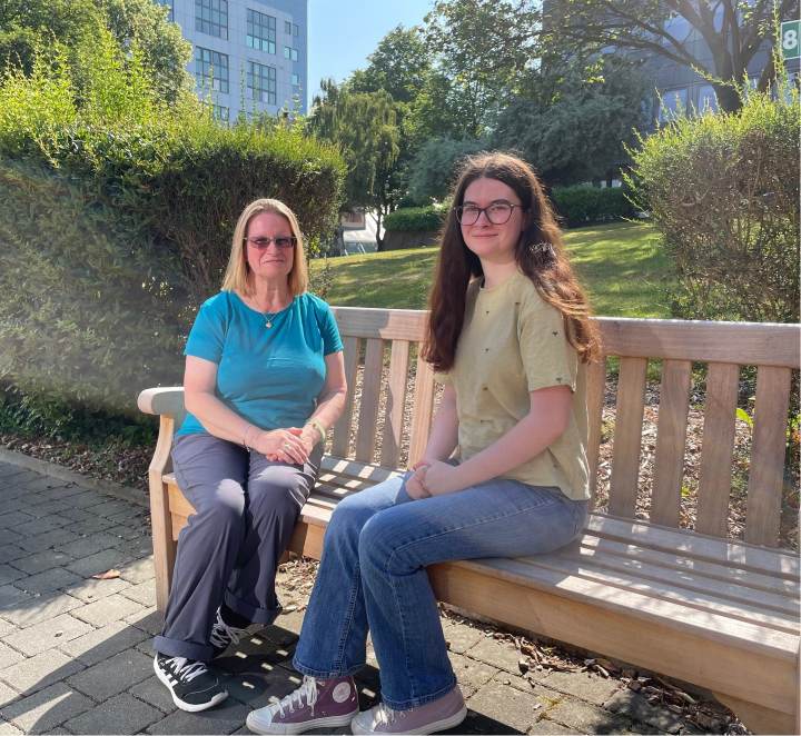 A mother and daughter sit down on a wooden bench with a university building behind them A mother and daughter sit down on a wooden bench with a university building behind them