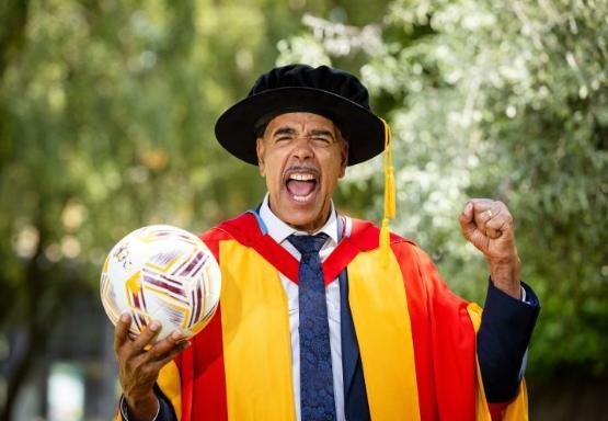 A person dressed in graduation gown holds a football in one hand and has a clinched fist in the other A person dressed in graduation gown holds a football in one hand and has a clinched fist in the other