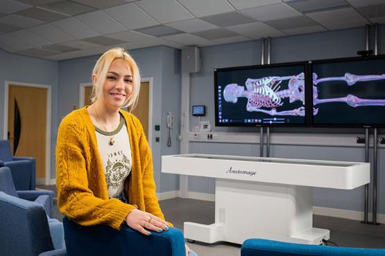 A postgraduate student sat in a chair in the Simulation Theatre. They are smiling at the camera.