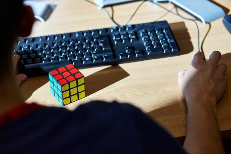 A person sat at a desk with a Rubik's cube on it, using a keyboard and mouse.